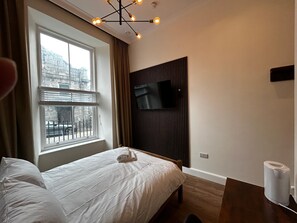 Ein Schlafzimmer mit einem Bett, einem an einer Holzplatte montierten Fernseher, einem Fenster mit Jalousien und einer Deckenleuchte.