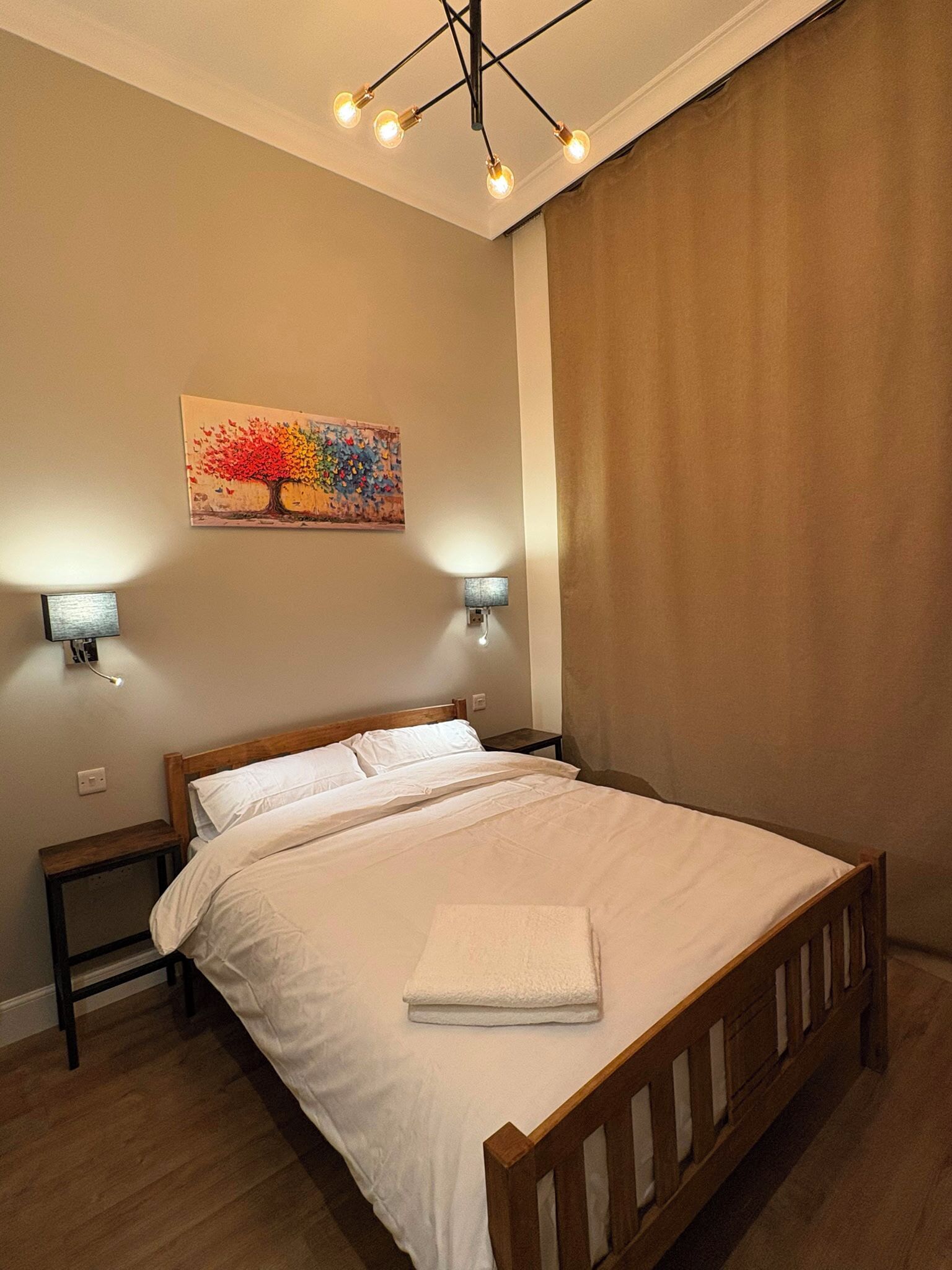 A bedroom with a wooden bed, two bedside tables, a wall-mounted painting, and a ceiling light fixture.