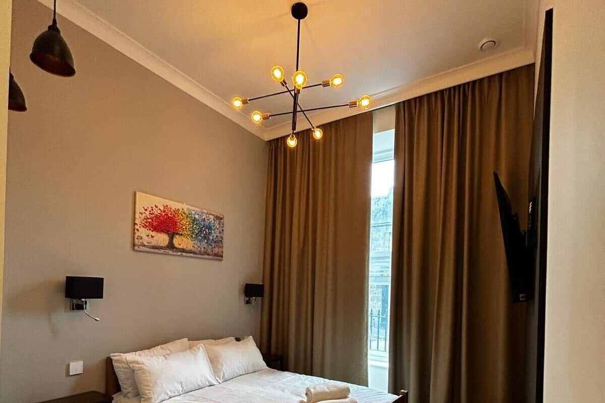 Un dormitorio con cama, escritorio, silla, un cuadro y ventana con cortinas.