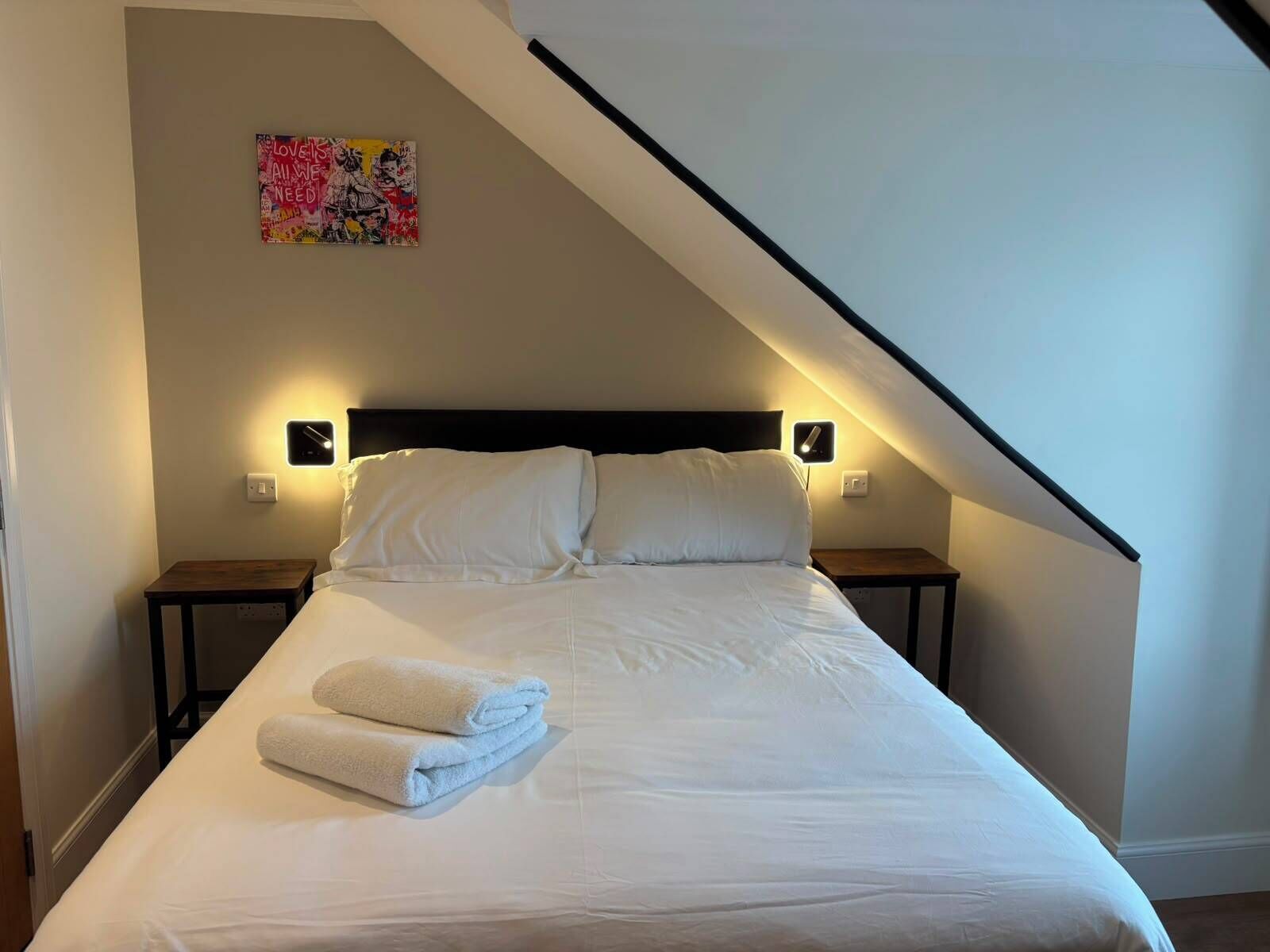 Ein ordentlich bezogenes Bett mit weißen Laken und einem gefalteten Handtuch, zwei Nachttische mit Lampen und ein farbenfrohes Kunstwerk an der Wand.