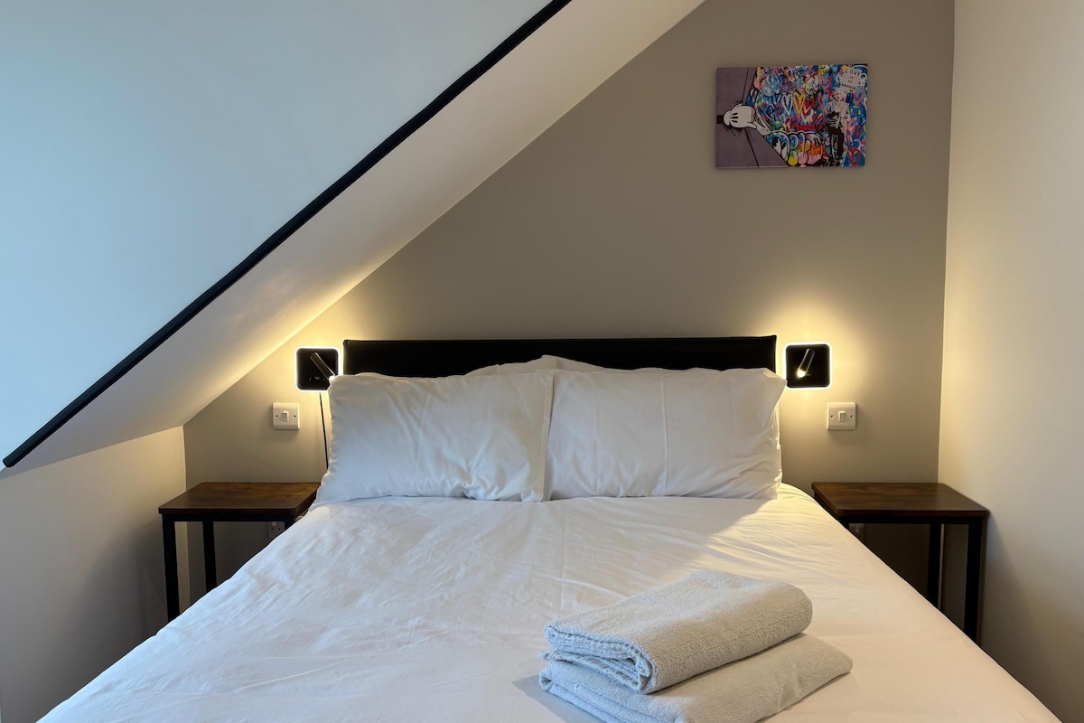 Un dormitorio con techo inclinado, una cama con ropa blanca, dos mesitas de noche y una lámpara de pared.