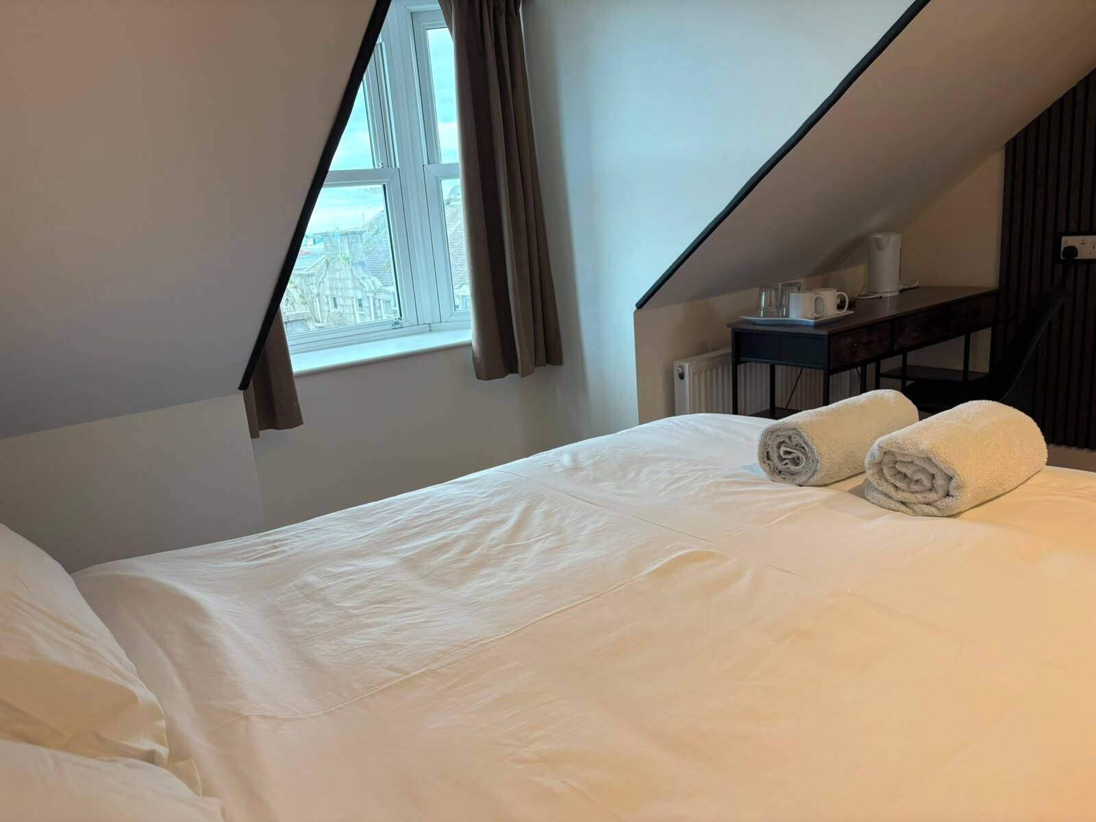 Una cama bien hecha con sábanas blancas y dos toallas enrolladas, una ventana con cortinas, y una mesita con una taza y un libro.