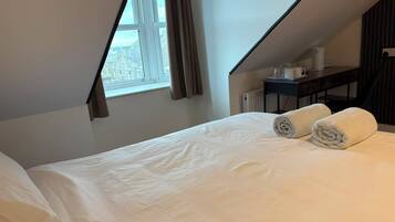 Una cama bien hecha con sábanas blancas y dos toallas enrolladas, una ventana con cortinas, y una mesita con una taza y un libro.