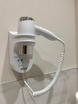 Un secador de pelo montado en la pared con cable, un panel de control con indicador rojo y una rejilla metálica.