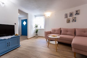 Ein Wohnzimmer mit Fernseher, einem rosafarbenen Sofa, einem kleinen runden Tisch, einer Zimmerpflanze und Bildern an der Wand.
