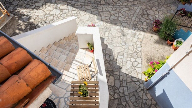 Um pátio com chão de pedra, um banco de madeira, plantas em vasos e uma parede branca.