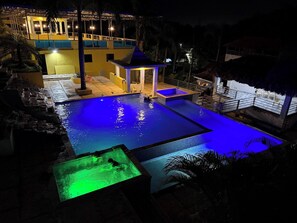 Ein Schwimmbecken mit einem kleinen angrenzenden Whirlpool, einer überdachten Sitzecke und einem Gebäude mit Balkon im Hintergrund.