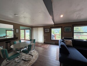 Ein Wohnzimmer mit Esstisch, Stühlen, einer Couch und großen Fenstern mit Blick auf Bäume.