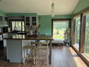 Une cuisine moderne dotée d’un grand îlot central, de tabourets en bois et offrant une vue sur une forêt par les fenêtres.