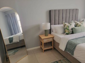 A bedroom with a bed, a bedside table with a lamp, a mirror, and a window with curtains. - Charming 2-Bedroom Apartment in Midrand, Close to Mall of Africa (Midrand)