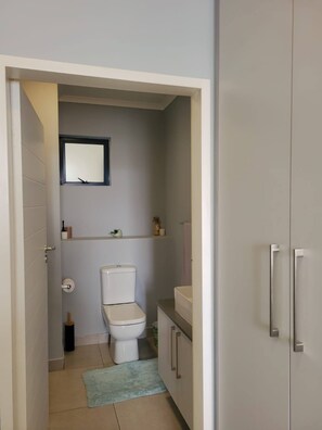 A compact bathroom with a toilet, a small shelf with toiletries, and a mirror above the toilet. - Charming 2-Bedroom Apartment in Midrand, Close to Mall of Africa (Midrand)