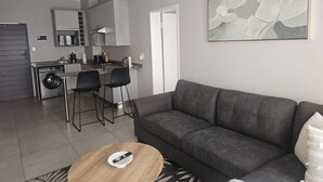 Un salon moderne avec un canapé gris, une table basse et un coin cuisine équipé d’un lave-linge et de tabourets de bar.