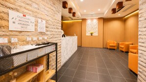 Zona de recepción con pared de piedra, mostrador de recepción y área de descanso con sillas naranjas.