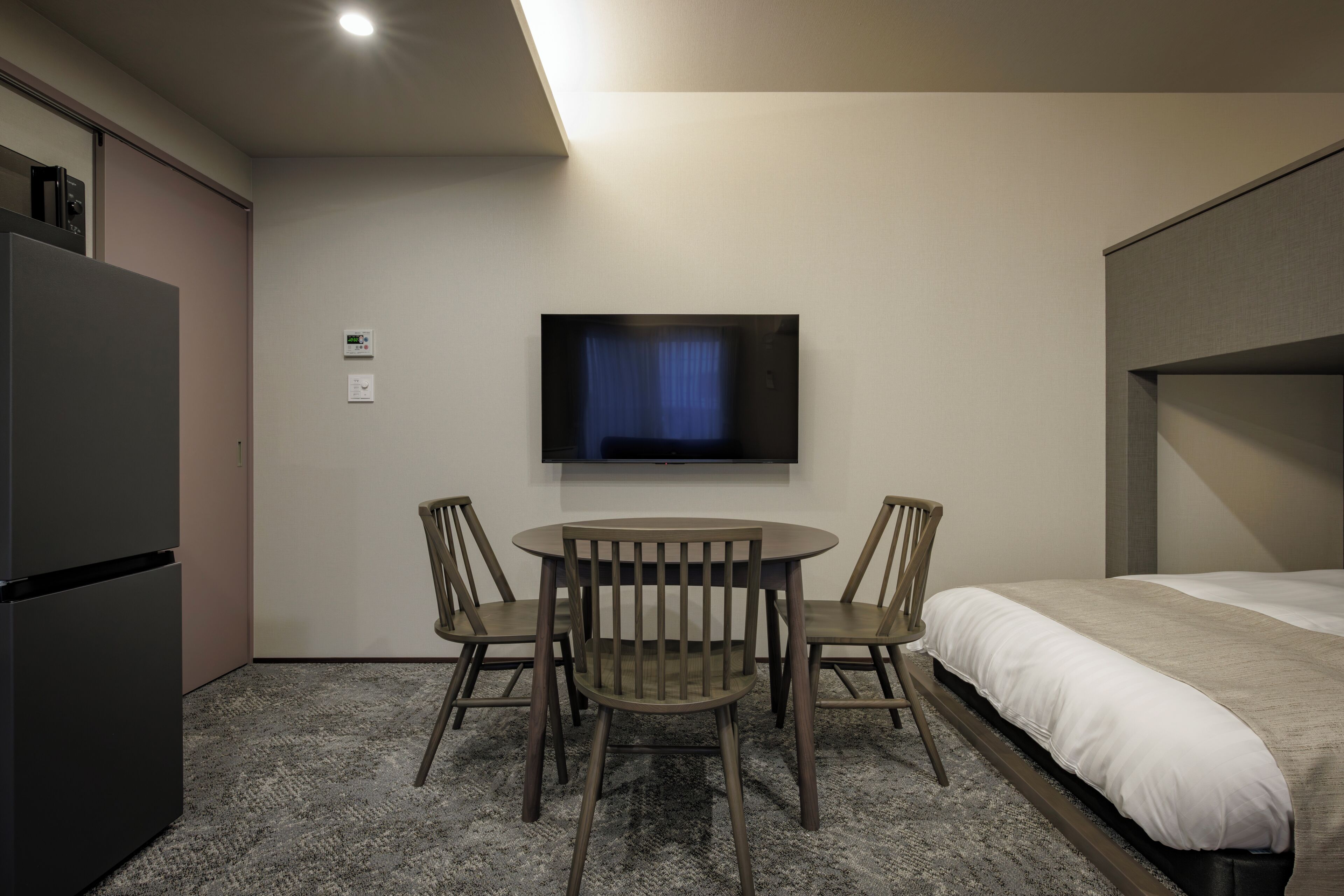 A hotel room with a bed, a TV mounted on the wall, a small table with chairs, and a refrigerator.