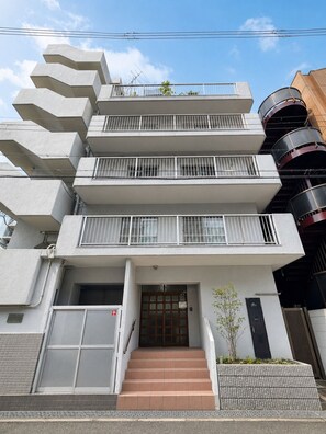 Ein mehrstöckiges Gebäude im modernen Stil, mit einem zentralen Eingang, einer Glastür, einer kleinen Außenfläche mit einer Pflanze und einer Treppe zum Eingang.