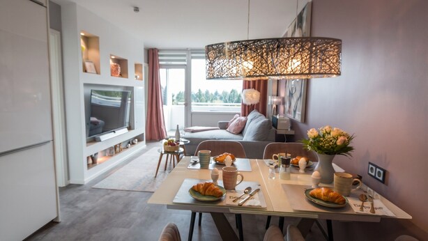 Et moderne spiseområde med et bord dækket til morgenmad, et fjernsyn i baggrunden og udsigt til det fri.