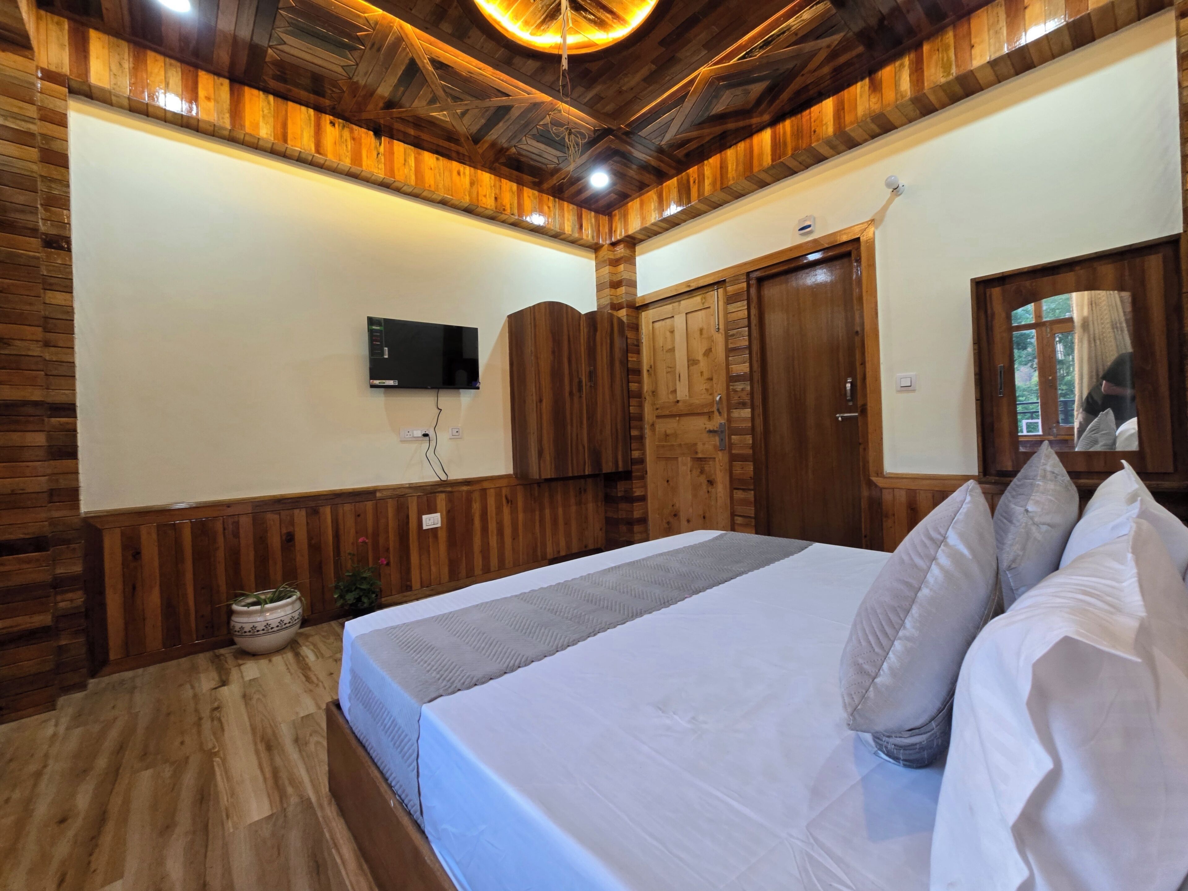 A bedroom with a wooden ceiling, a bed with pillows, a television on the wall, and a wooden wardrobe.