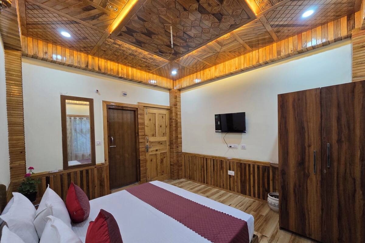 A bedroom with a wooden ceiling, a bed with white and red bedding, a TV mounted on the wall, and wooden wardrobes.
