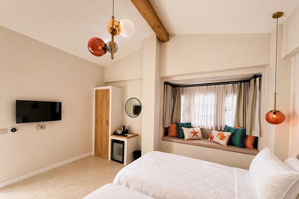 Un dormitorio con televisor, armario de madera, mesita de noche pequeña, asiento junto a la ventana con cojines e iluminación colgante.