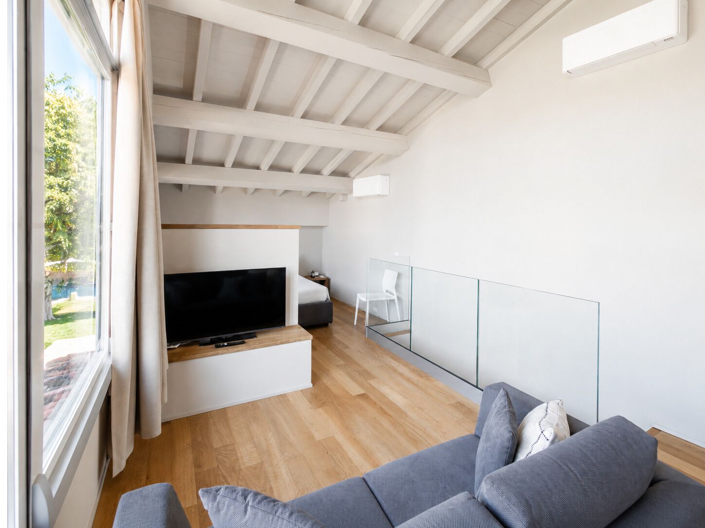 A modern living room with a flat-screen TV, a grey sofa, and a glass partition.