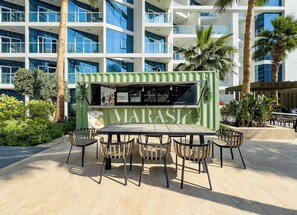 Un espacio de comedor al aire libre MARASI con un quiosco verde, rodeado de palmeras y edificios modernos.