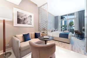Habitación de hotel con cama, sofá, mesa de comedor, una silla, lámpara y un gran ventanal con vistas a edificios.