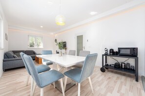 A modern kitchen with a white table, blue chairs, a grey sofa, and a black shelf with kitchen appliances. - Pool House Olive (Zemunik Donji)