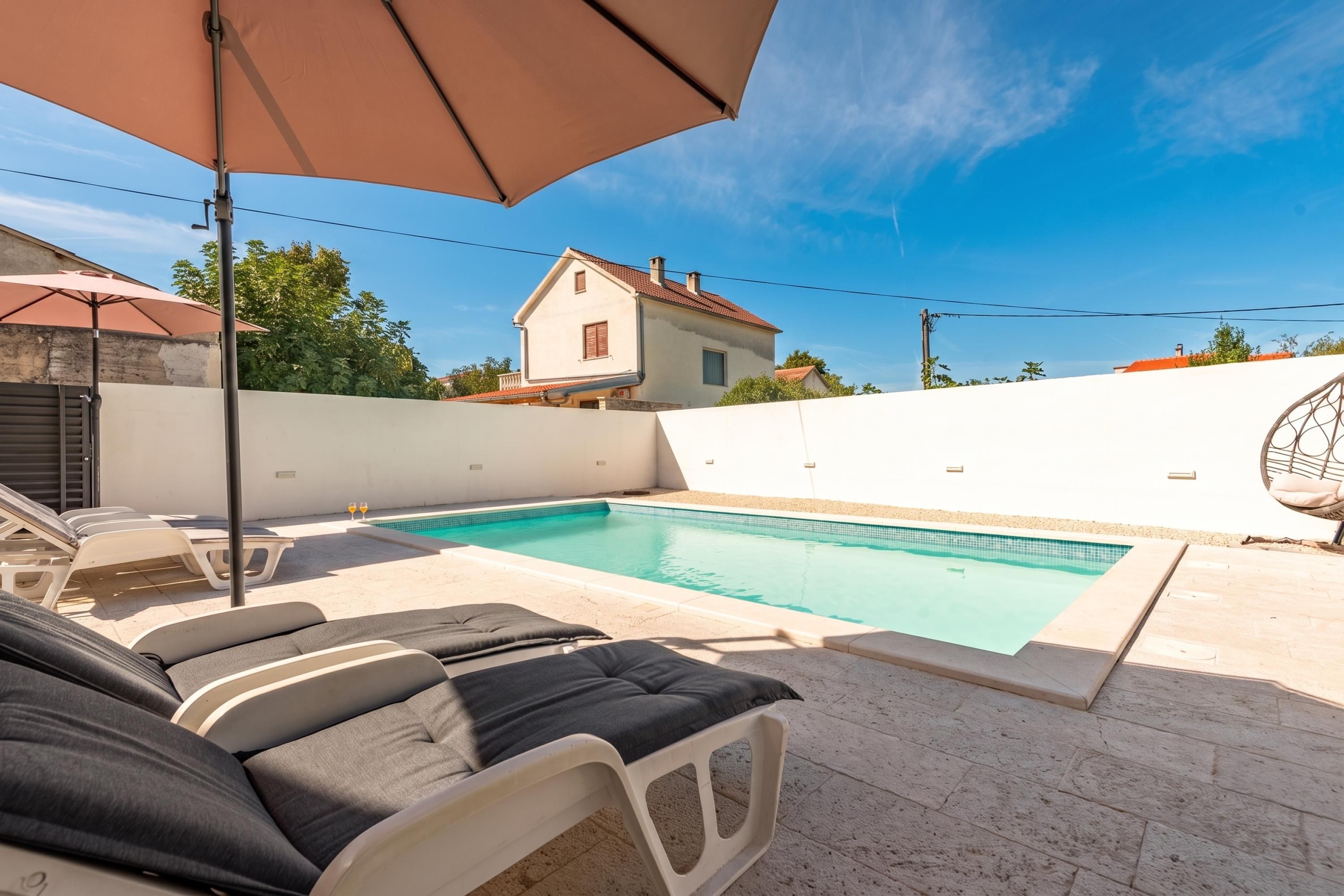Une piscine entourée de chaises longues et dotée d’un parasol, le tout bordé d’un mur blanc et avec une maison en arrière-plan.