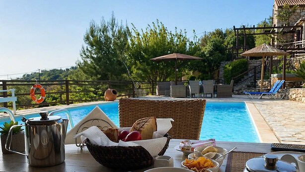 A dining table with breakfast items, a metal teapot, and a cup, overlooking a swimming pool with lounge chairs and a stone building in the background. - Villa Poseidon - Master Suite In Zante w/Pool (Skinaria)