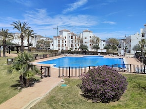 A swimming pool surrounded by a fence, palm trees, and a building complex in the background. - Family Golf Oasis | Disney+, Pool & Sunny Terrace (Murcia)