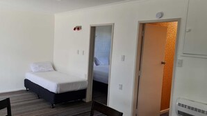 A small, single-bed room with a wooden floor, a chair, a door, and a wall-mounted heater.