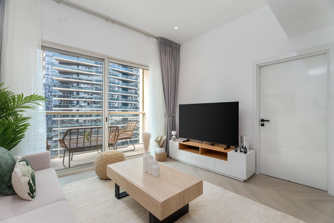 A modern living room with a large TV, a sofa, a coffee table, and a balcony with a view of high-rise buildings.