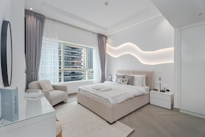 A modern bedroom with a large bed, a sitting area, and a view of high-rise buildings. - Gala Guests - Marina Views Collection (Dubai)