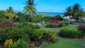 A tropical garden with palm trees and colorful flowers, a grassy lawn, and a view of the ocean. - Castles in Paradise #C5 - by ZenBreak (Savannes)