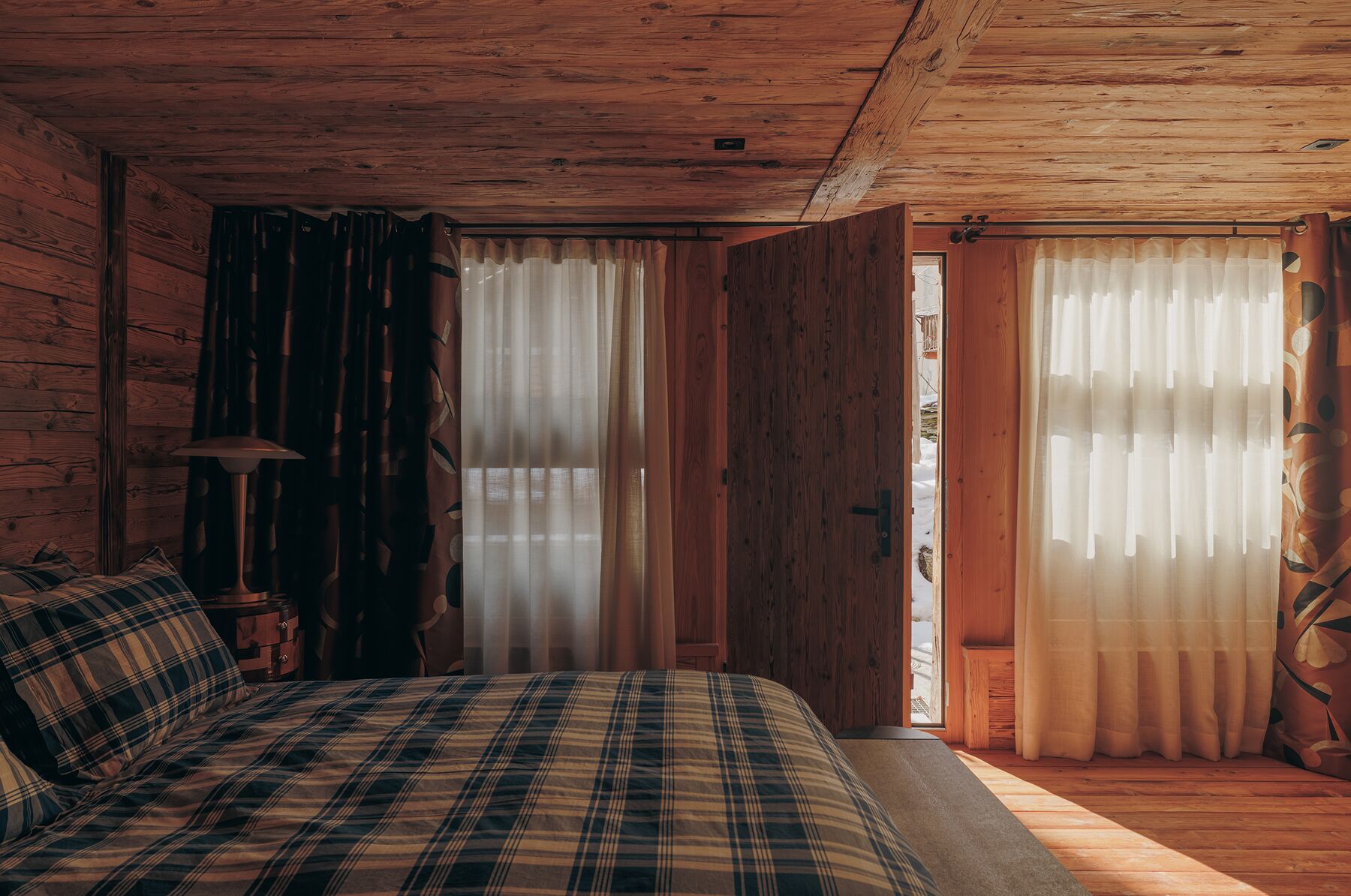 Ein Schlafzimmer einer Holzhütte mit einer Karo-Bettdecke, einem Nachttisch mit Lampe und zwei Fenstern mit durchsichtigen Vorhängen.