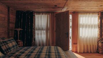 Ein Schlafzimmer einer Holzhütte mit einer Karo-Bettdecke, einem Nachttisch mit Lampe und zwei Fenstern mit durchsichtigen Vorhängen.