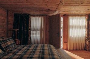 A wooden cabin bedroom with a plaid bedspread, a nightstand with a lamp, and two windows with sheer curtains.
