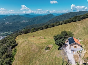 A house with a sloped roof, surrounded by a grassy field and trees, with a mountain range in the background.