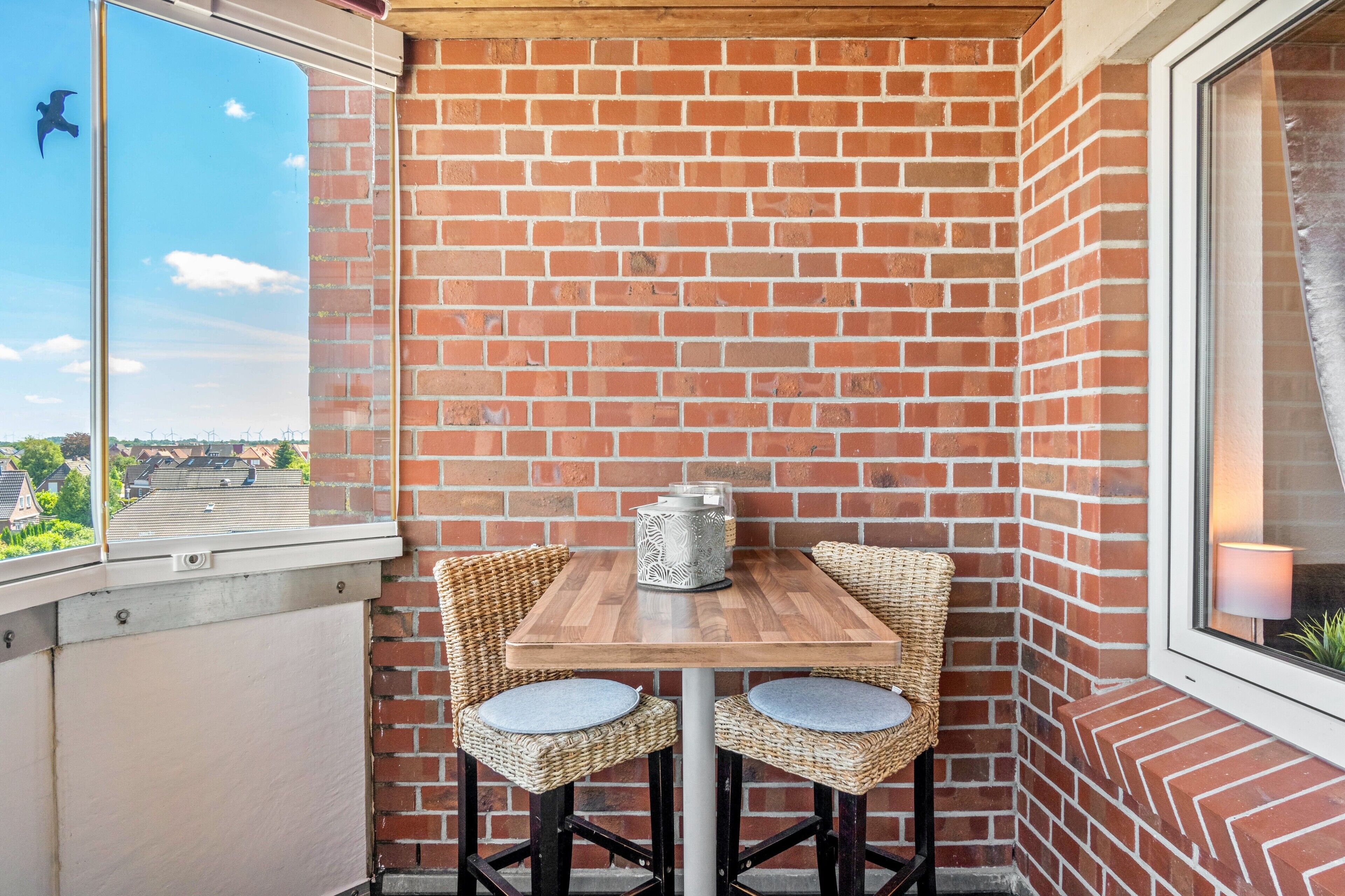 Een hoekbalkon met een bakstenen muur, een houten tafel met twee rieten stoelen en uitzicht op windmolens.