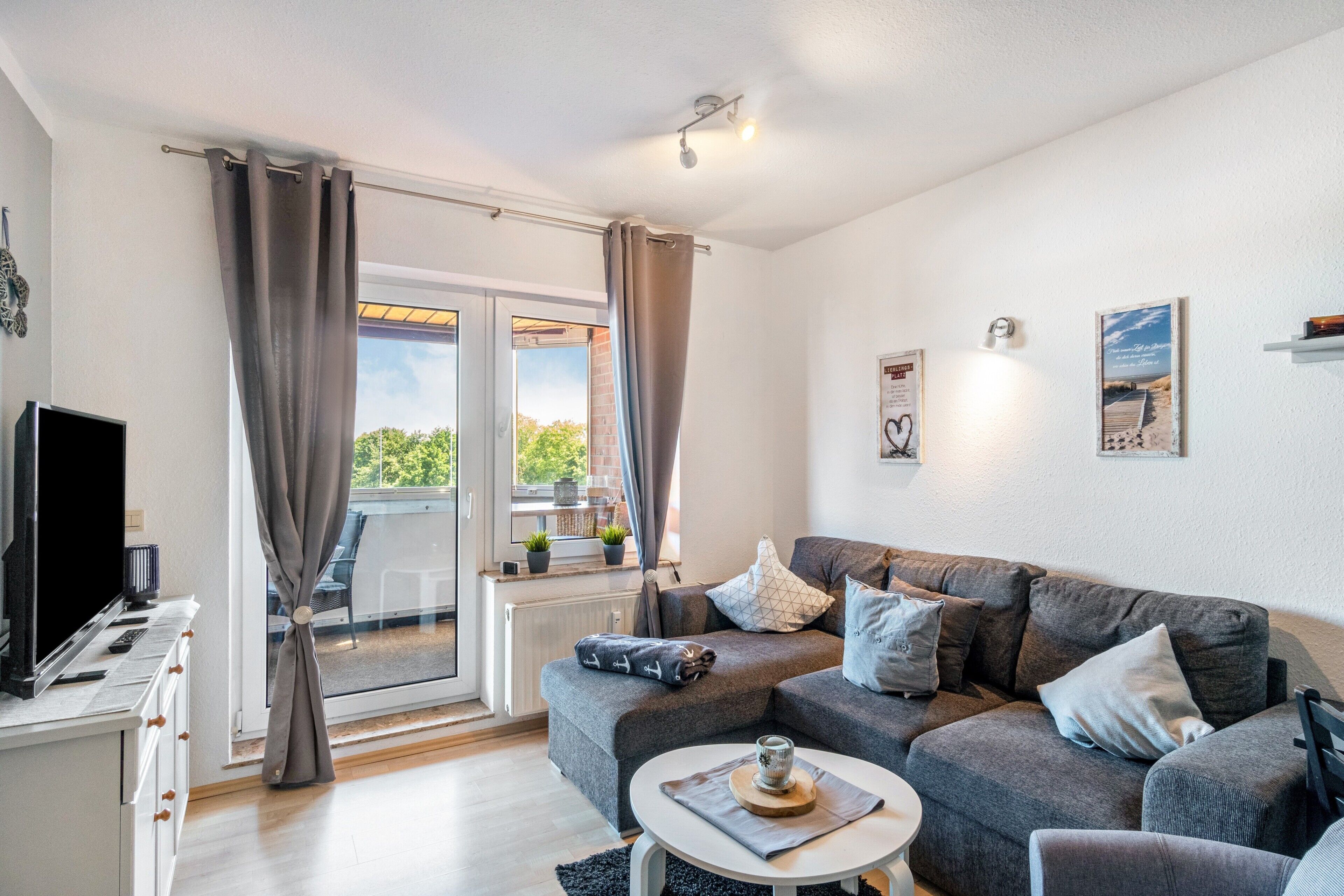 Ein modernes Wohnzimmer mit grauem Sofa, einem Couchtisch, einem Fernseher und einem Balkon mit Vorhängen.