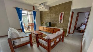 Ein Wohnzimmer mit einer Couch, einem Holztisch und einem Fernseher. Es gibt einen Essbereich mit Stühlen und einem Tisch. Der Raum verfügt über ein großes Fenster mit Vorhängen, eine Wand-Klimaanlage und ein Gemälde.
