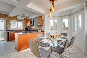 A modern kitchen with a central island, a dining area with a glass table, and large windows. - Kalisti-4 Bedroom Beachfront Villa with Private Pool (Sotira Ammochostou)