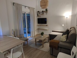 Ein modernes Wohnzimmer mit Esstisch, Sofa, Fernseher und einem großen Fenster mit Vorhängen.