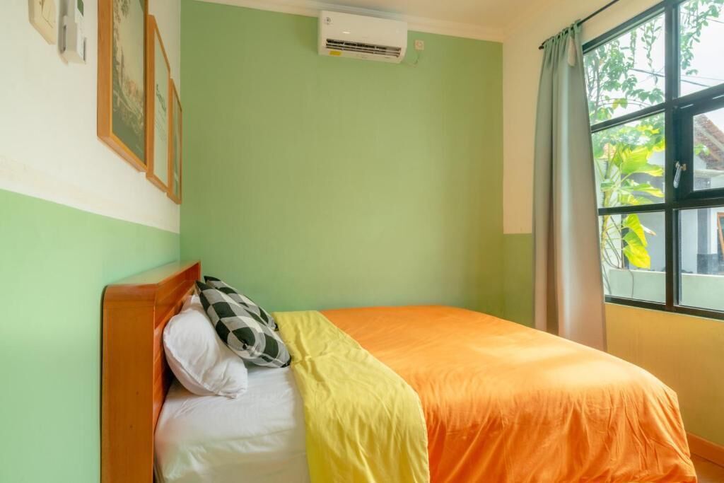 A single bed with a wooden headboard, a white pillow, a checkered pillow, and an orange and yellow bedspread. There is an air conditioning unit on the wall, a window with a curtain, and a green wall.