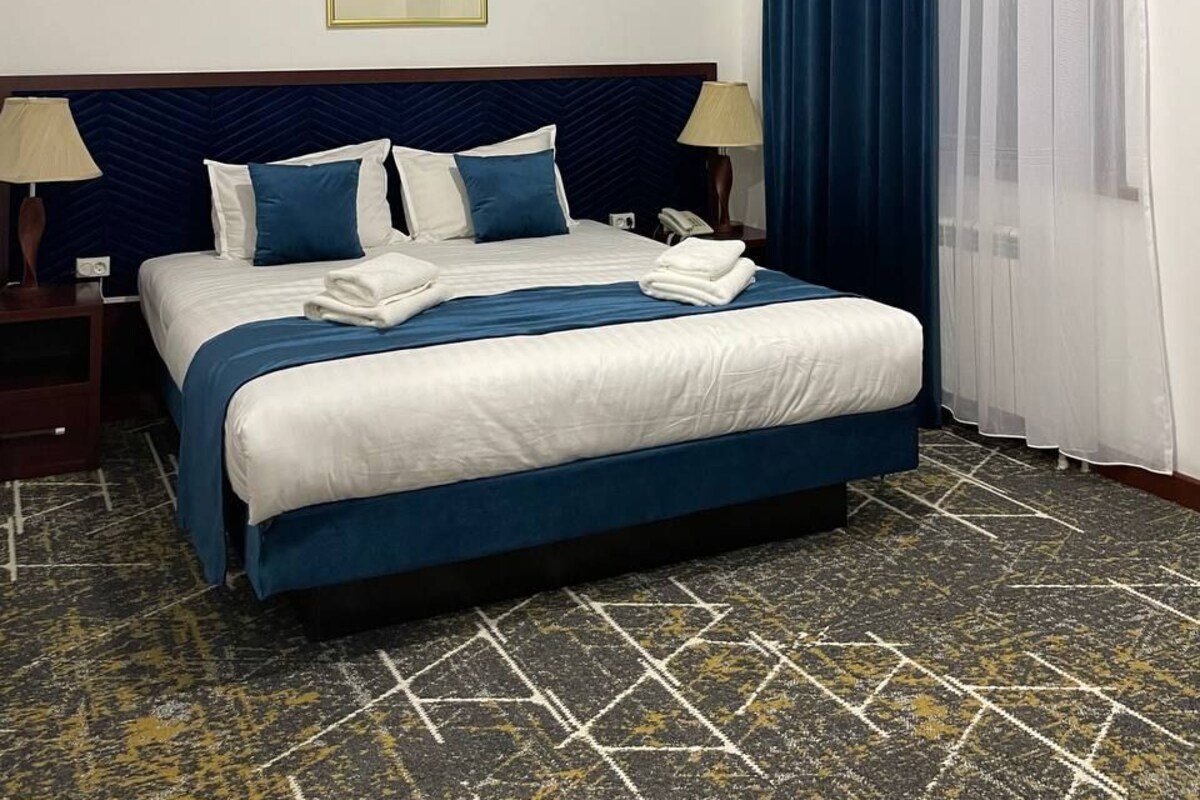 A hotel room with a bed, two bedside lamps, a window with blue curtains, and a wall-mounted picture.