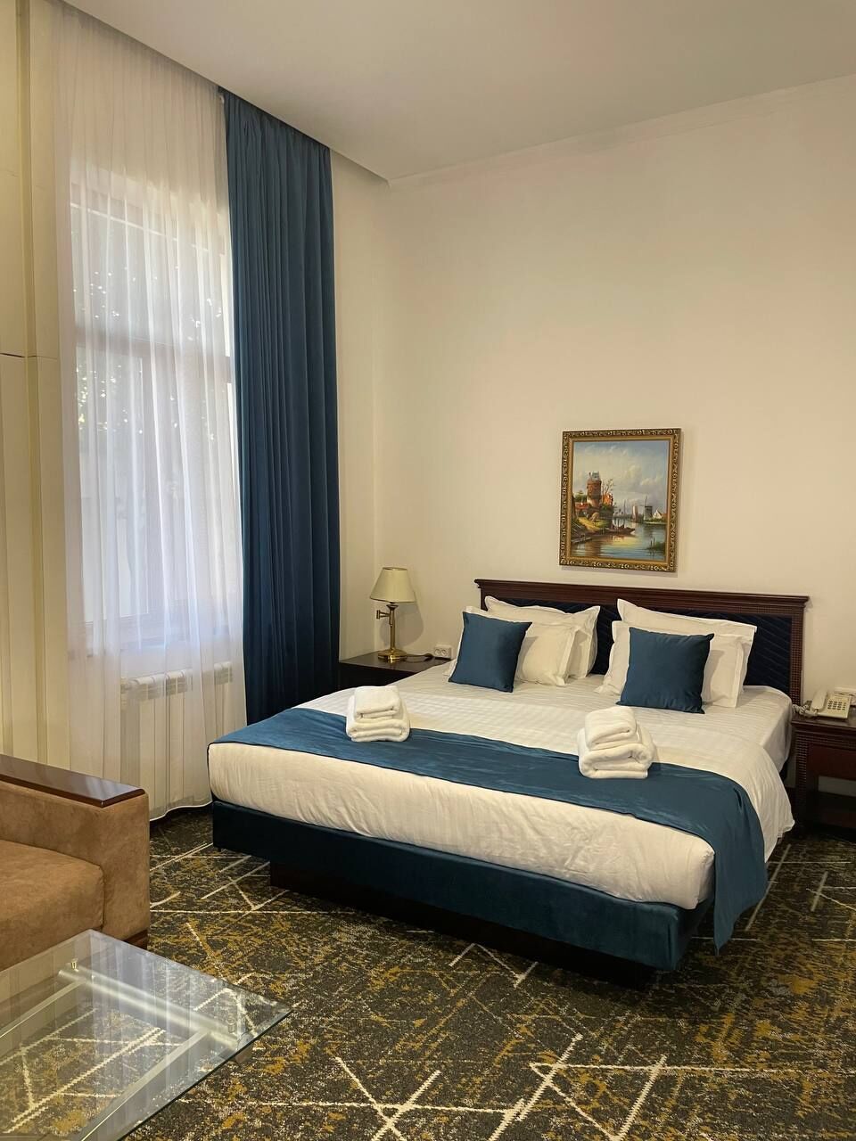 A hotel room with a bed, a nightstand, a lamp, a painting, and a patterned carpet.