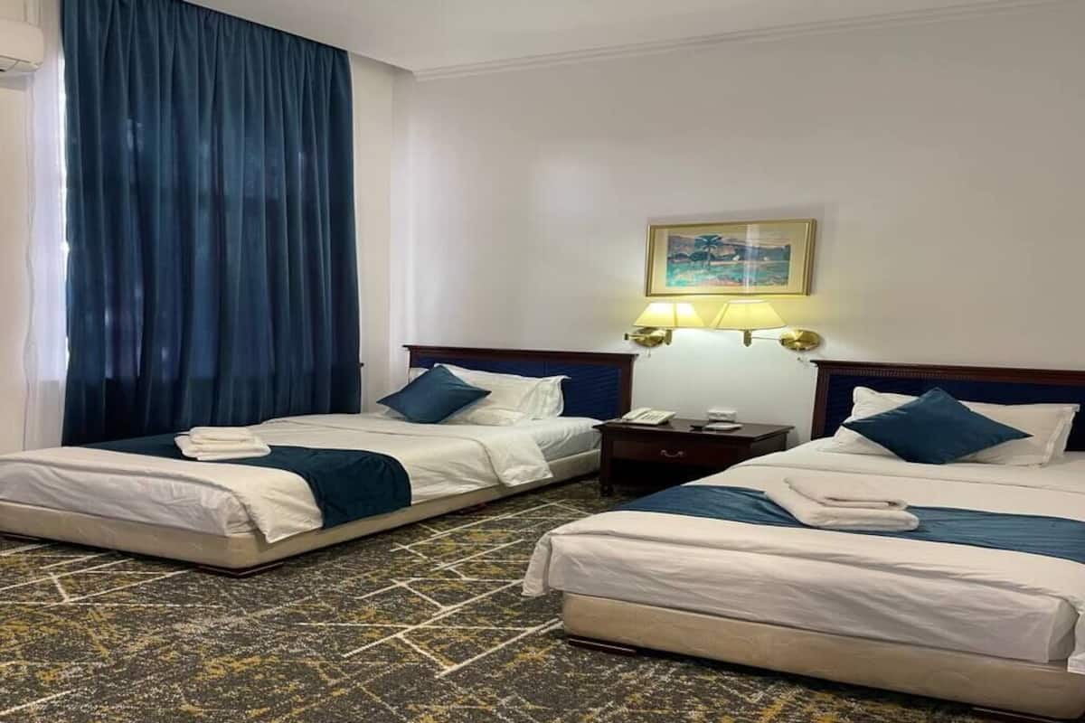 A hotel room with two beds, a large window with curtains, a painting on the wall, and a nightstand with a lamp.
