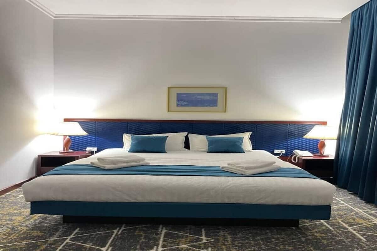A hotel room with a large bed, two bedside tables, a framed picture on the wall, and blue curtains.