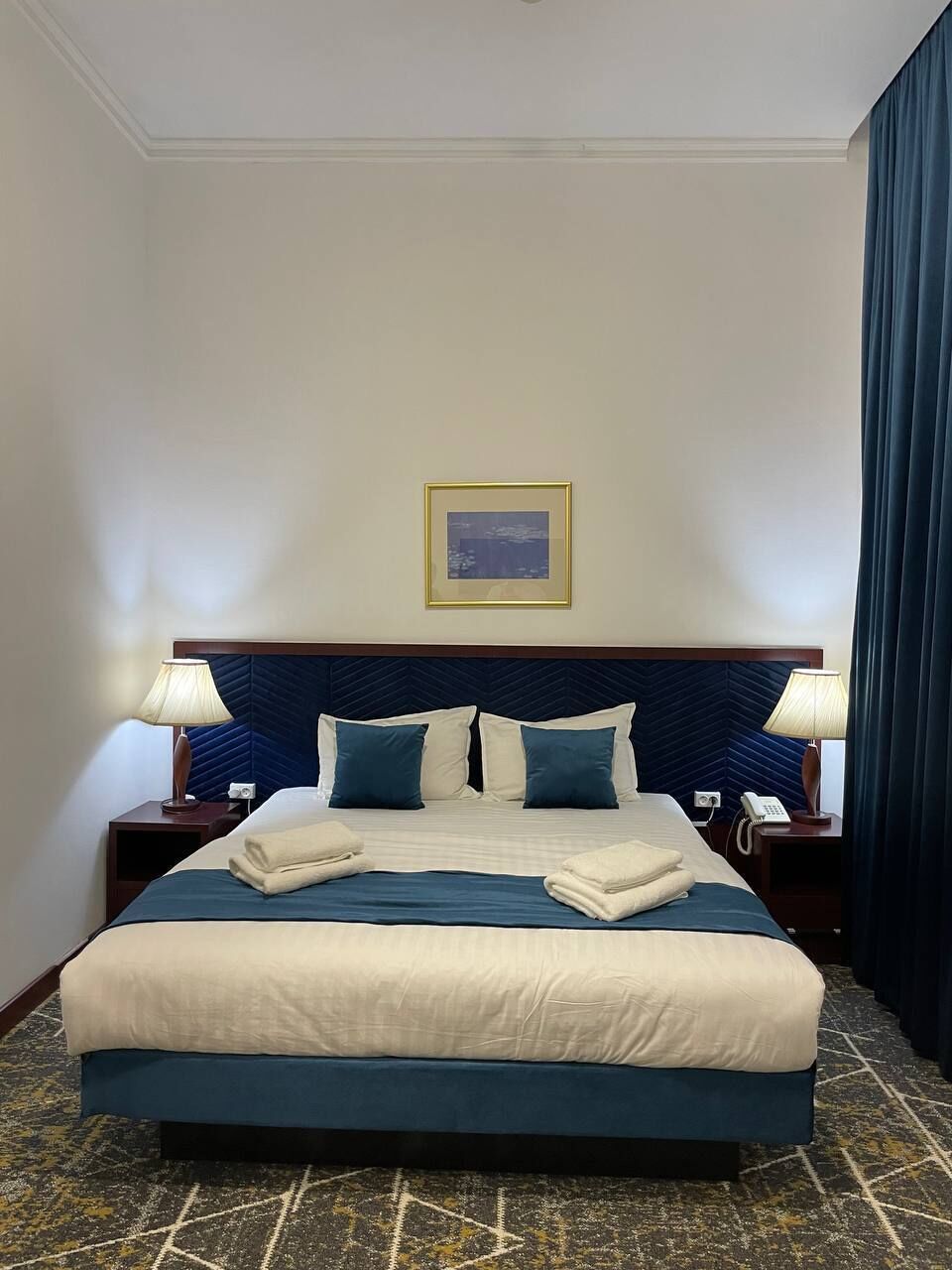 A hotel room with a large bed, two bedside tables, a headboard, a framed picture on the wall, and a patterned carpet.
