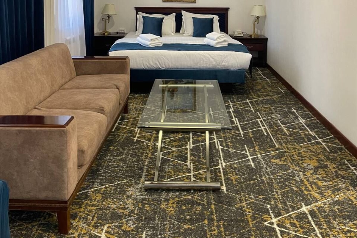 A hotel room with a bed, a sofa, a glass coffee table, and a painting on the wall.